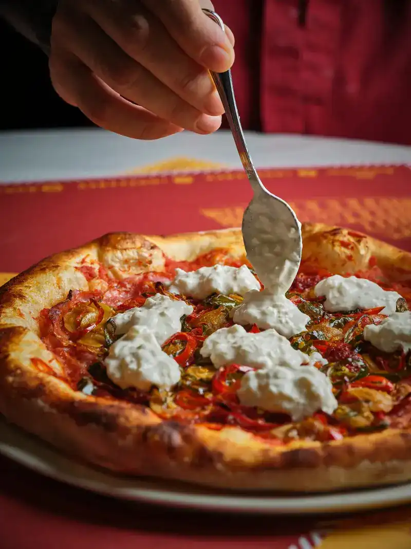 A spoon dropping creamy white cheese onto a pizza with a blistered crust