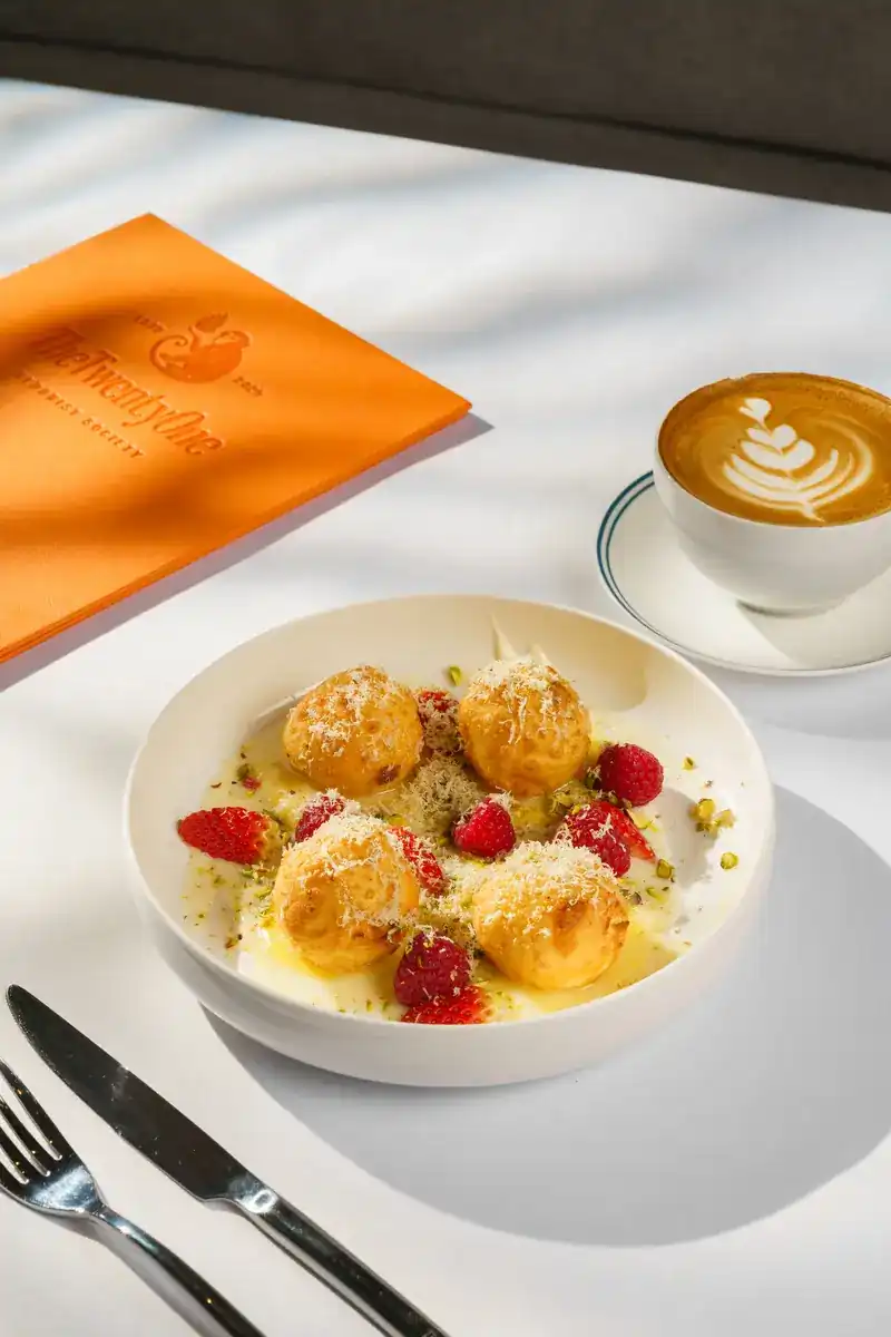 Golden pastry puffs with raspberries and pistachio, a latte and the orange menu cover on a white tablecloth