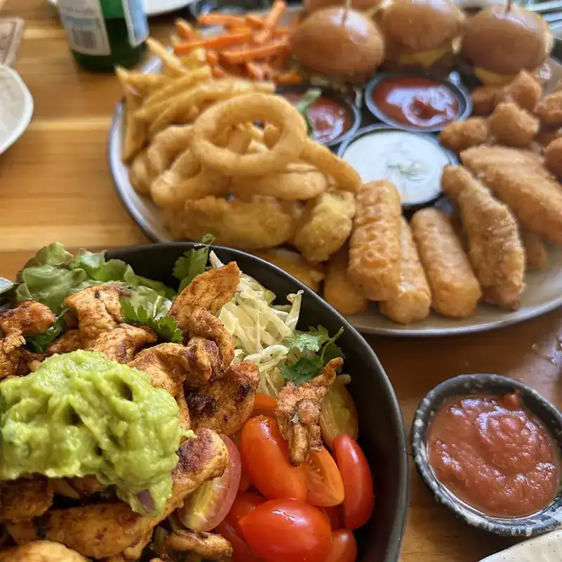 A sampler platter of mini burgers, onion rings and mozzarella sticks beside a bowl with chicken and guacamole