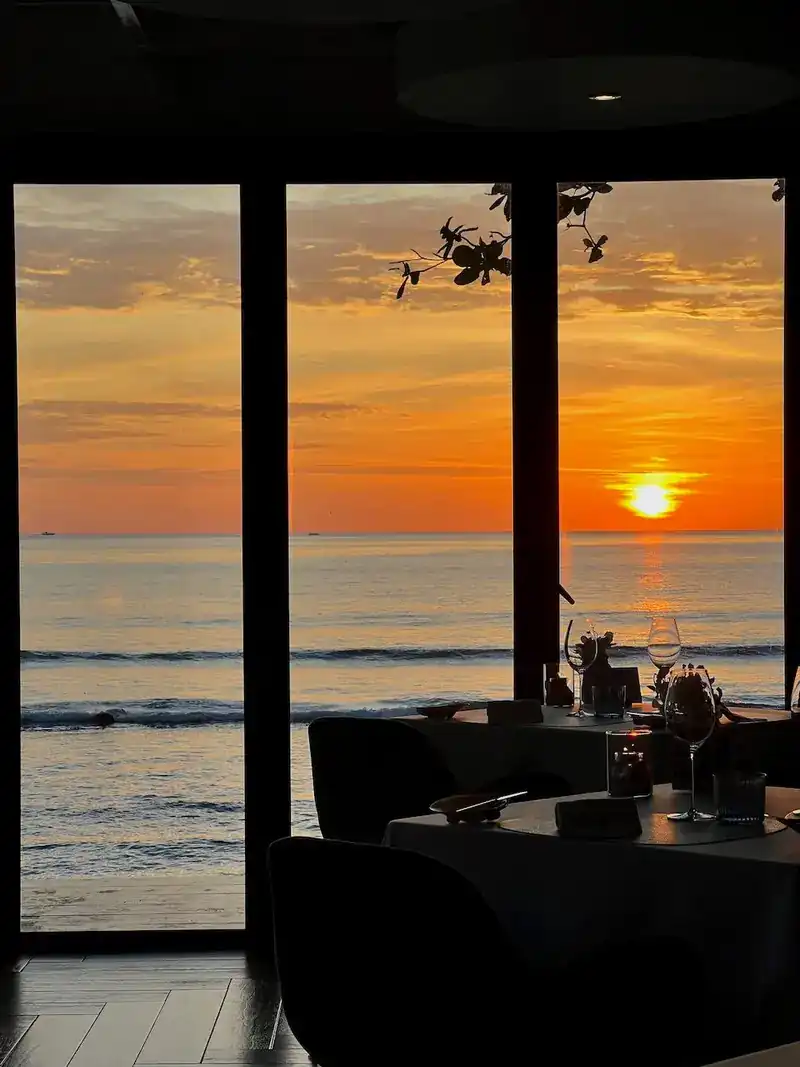 The dining room at sunset, tables set beside tall windows facing the sea
