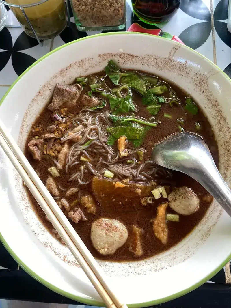Boat noodles in dark broth with meatballs, sliced meat and greens