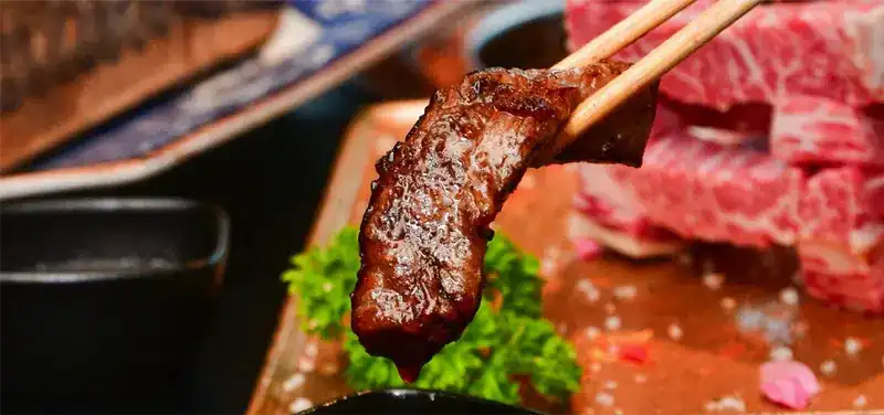 A piece of grilled beef held in chopsticks over a dish of sauce