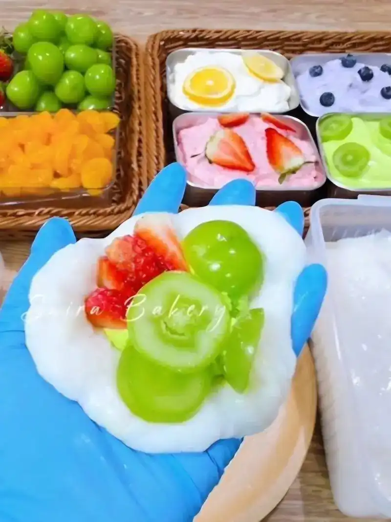A gloved hand holding soft white dough topped with strawberry and green grapes, trays of fruit and cream behind
