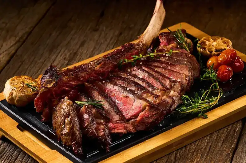 A sliced Black Angus tomahawk on a board with roasted garlic and tomatoes