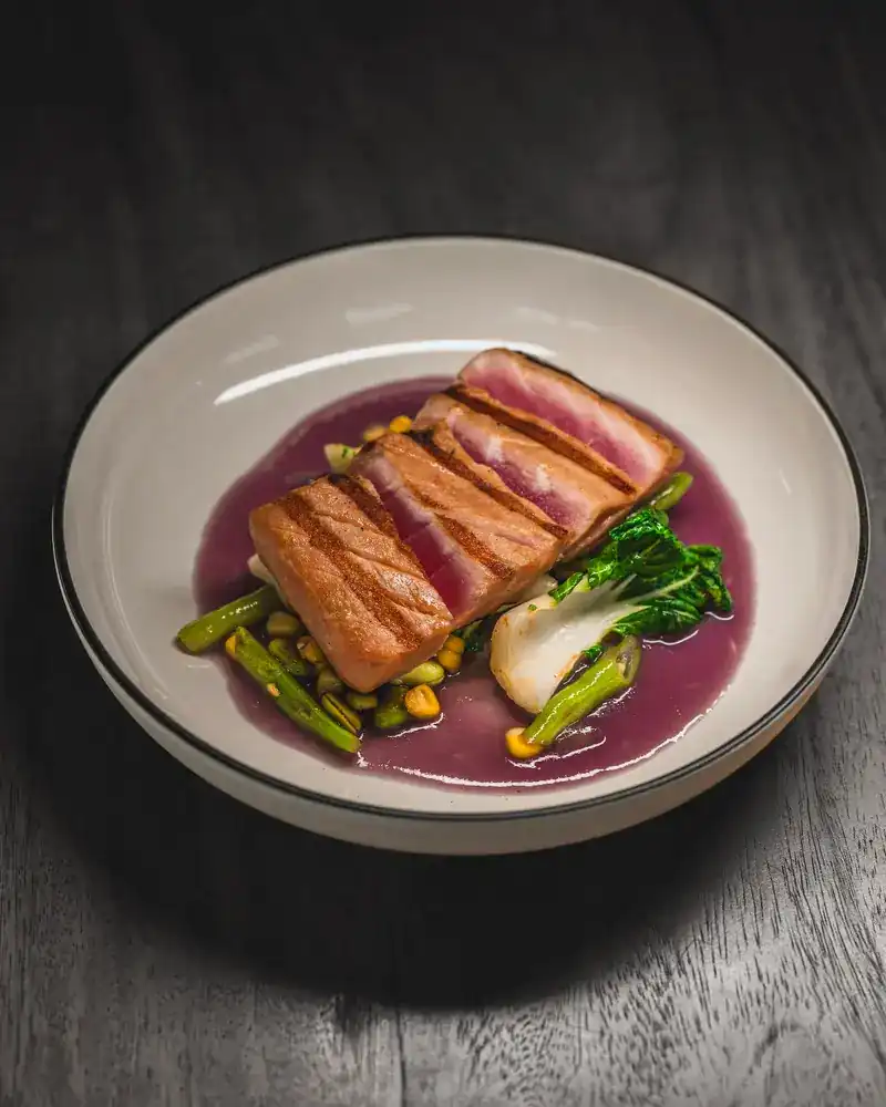 A seared fish fillet on a purple sauce with bok choy, asparagus and corn in a white bowl