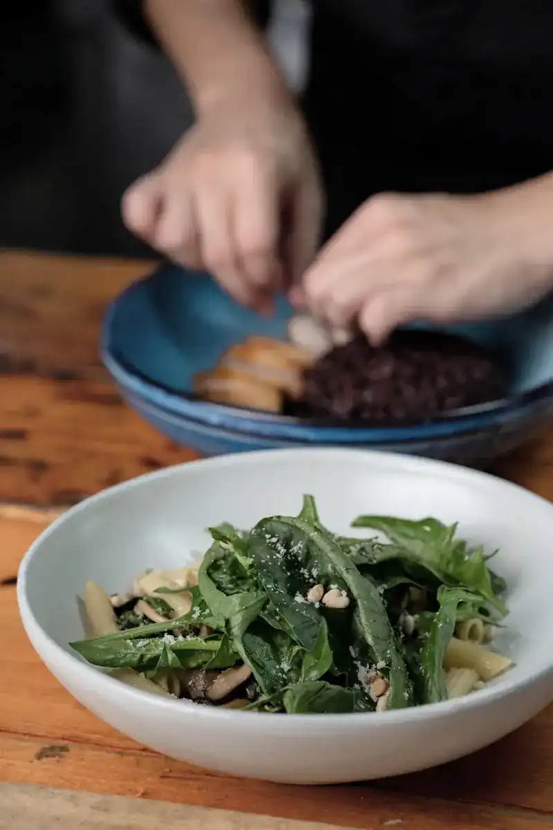 A bowl of penne with spinach and pine nuts, a cook's hands plating behind it