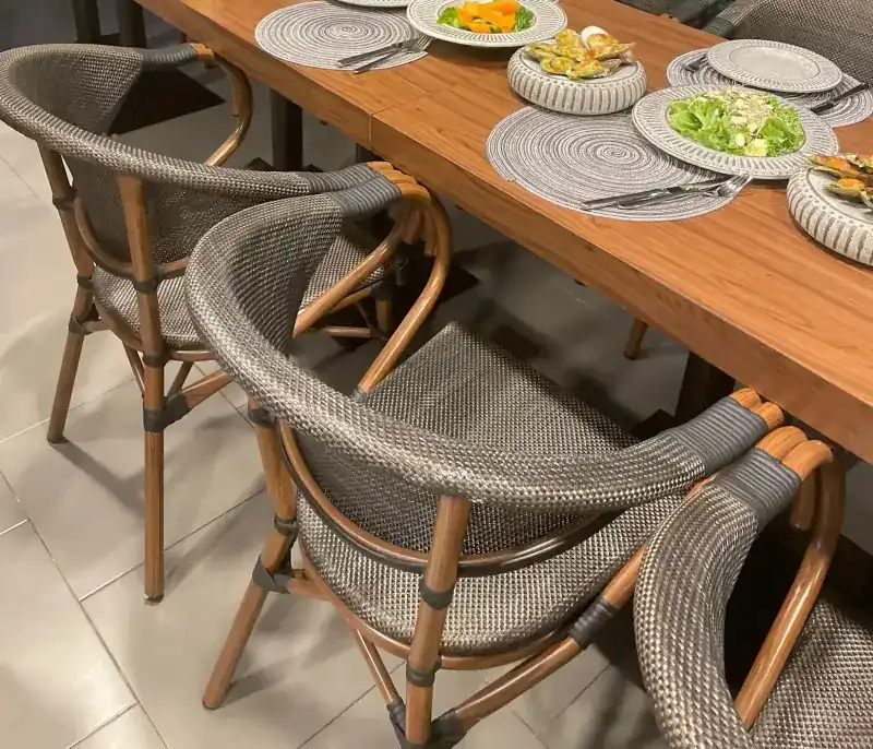 Woven rattan chairs around a long wooden table laid with ribbed grey plates of salad and baked mussels