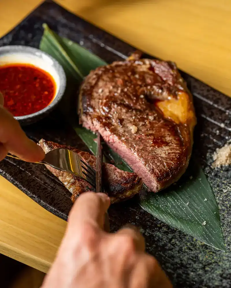 A steak being cut on a dark stone plate, with a chilli dip
