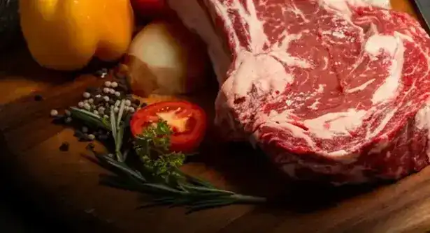 A raw dry-aged prime rib on a wooden board with peppercorns, rosemary and tomato