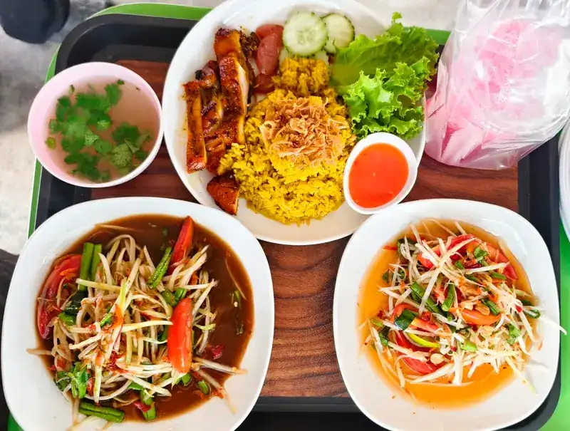 A tray with yellow khao mok rice and grilled chicken, two plates of som tam and a cup of soup