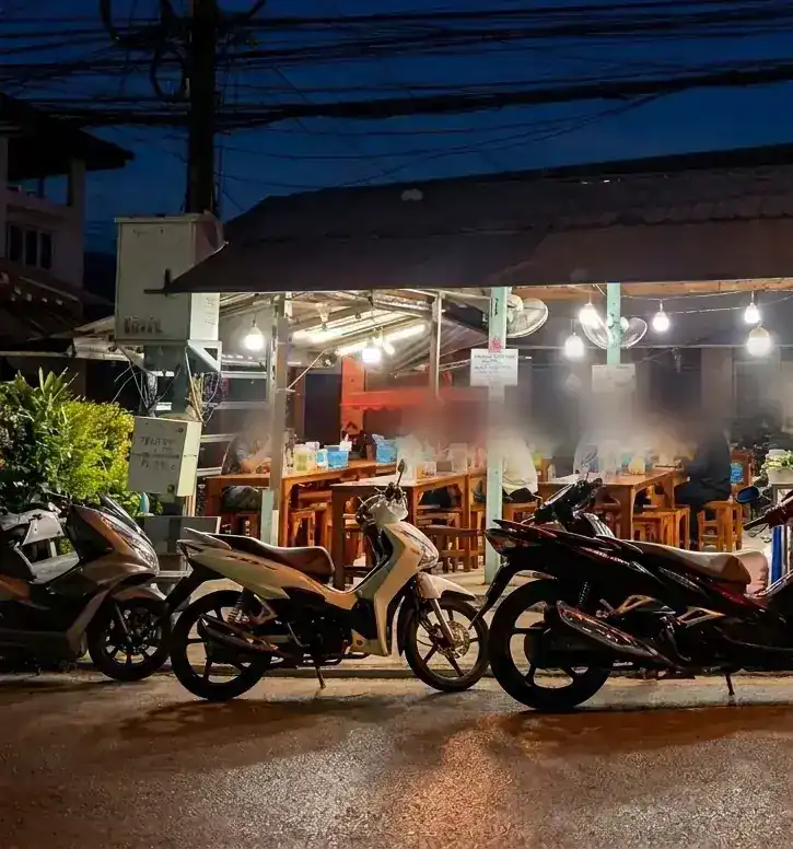 Evening at the roadside: lights under the roof, diners at wooden tables, motorbikes parked in front.