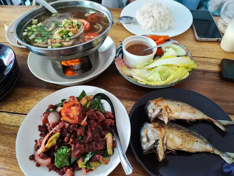 A soup in a hot pot over a flame, a red chilli stir-fry, two fried mackerel, rice, and vegetables with a dip