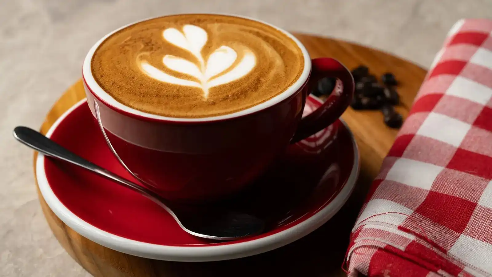 A latte with leaf latte art in a red cup, beside a red checked napkin