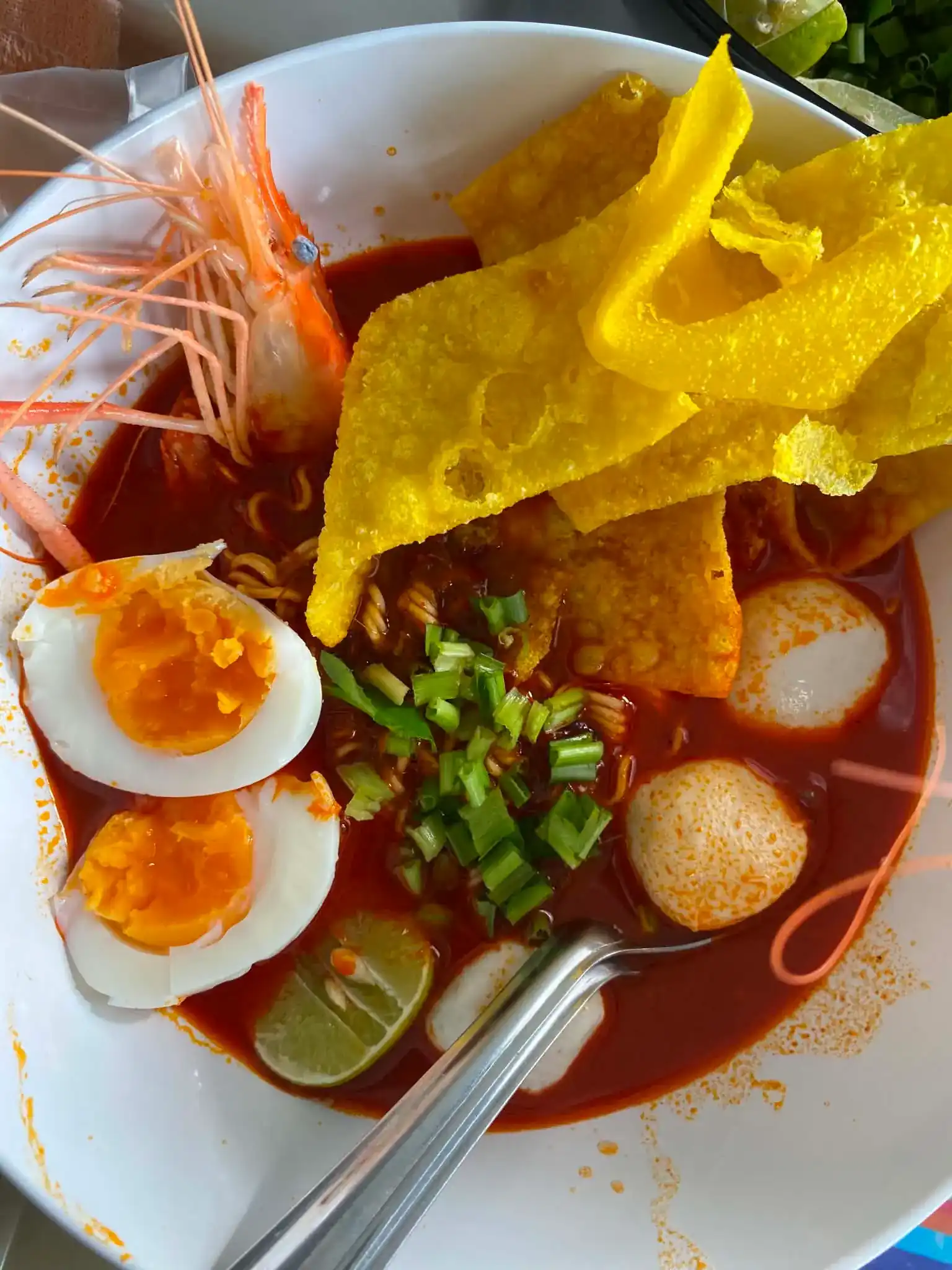 A bowl of tom yum noodles: river prawn, crisp fried wonton, halved boiled egg, lukchin, lime and spring onion in red broth