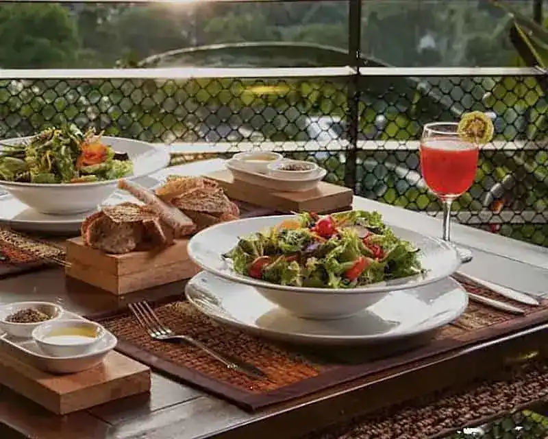 Two salads, sliced bread on a wooden board and a red juice on a table by the railing