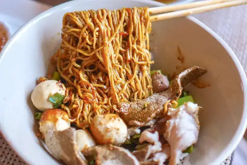 Egg noodles lifted from the bowl with chopsticks, with meatballs below