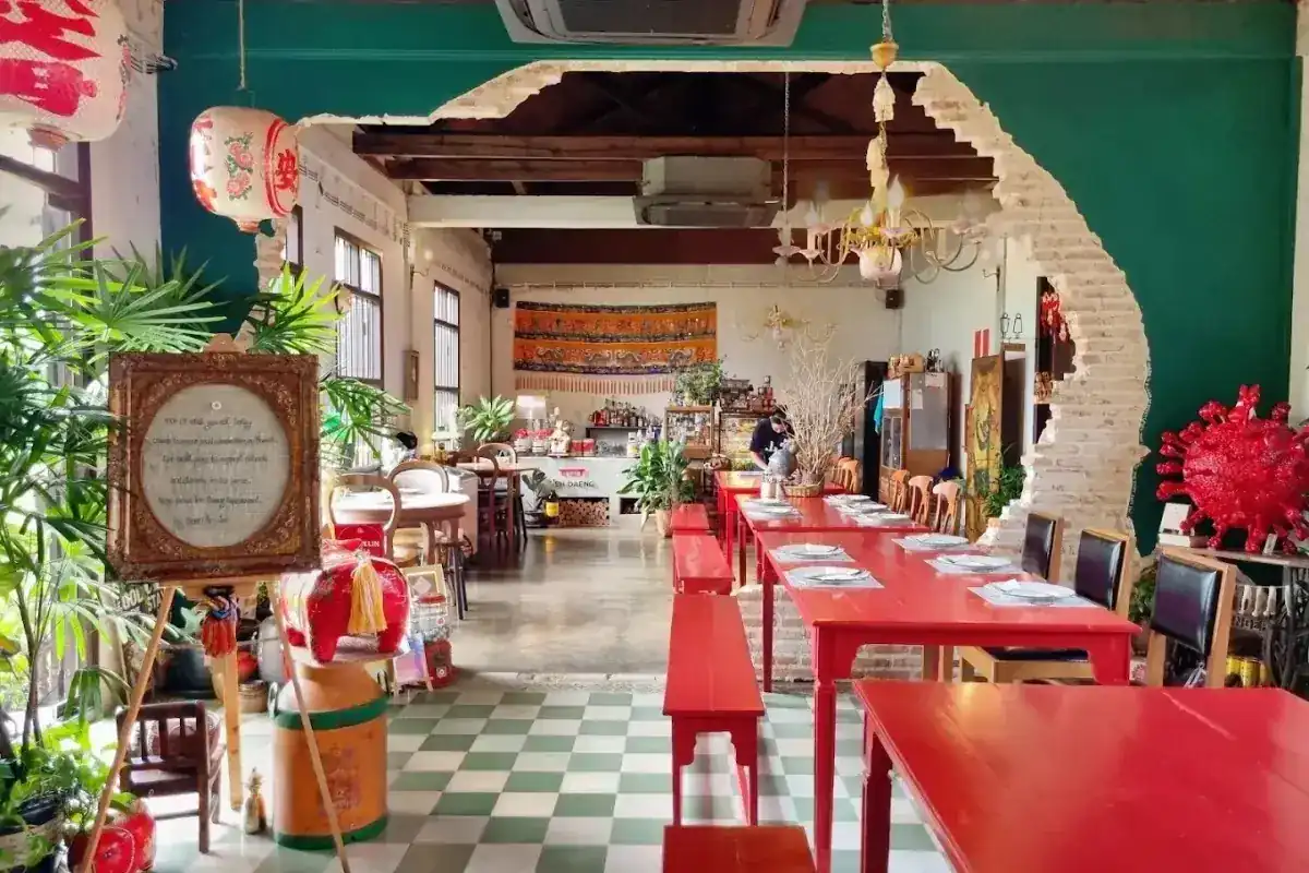 The dining room at Baan Ar-Jor: long red tables and benches on a green and white tiled floor, seen through a broken brick opening in a green wall