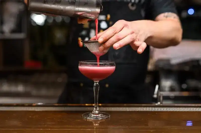 A bartender straining a pink cocktail into a coupe glass