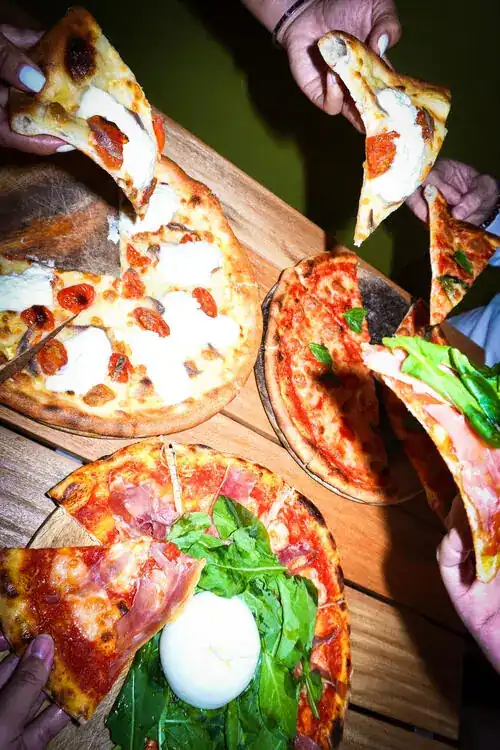 Friends' hands reaching for slices from several pizzas on a wooden table