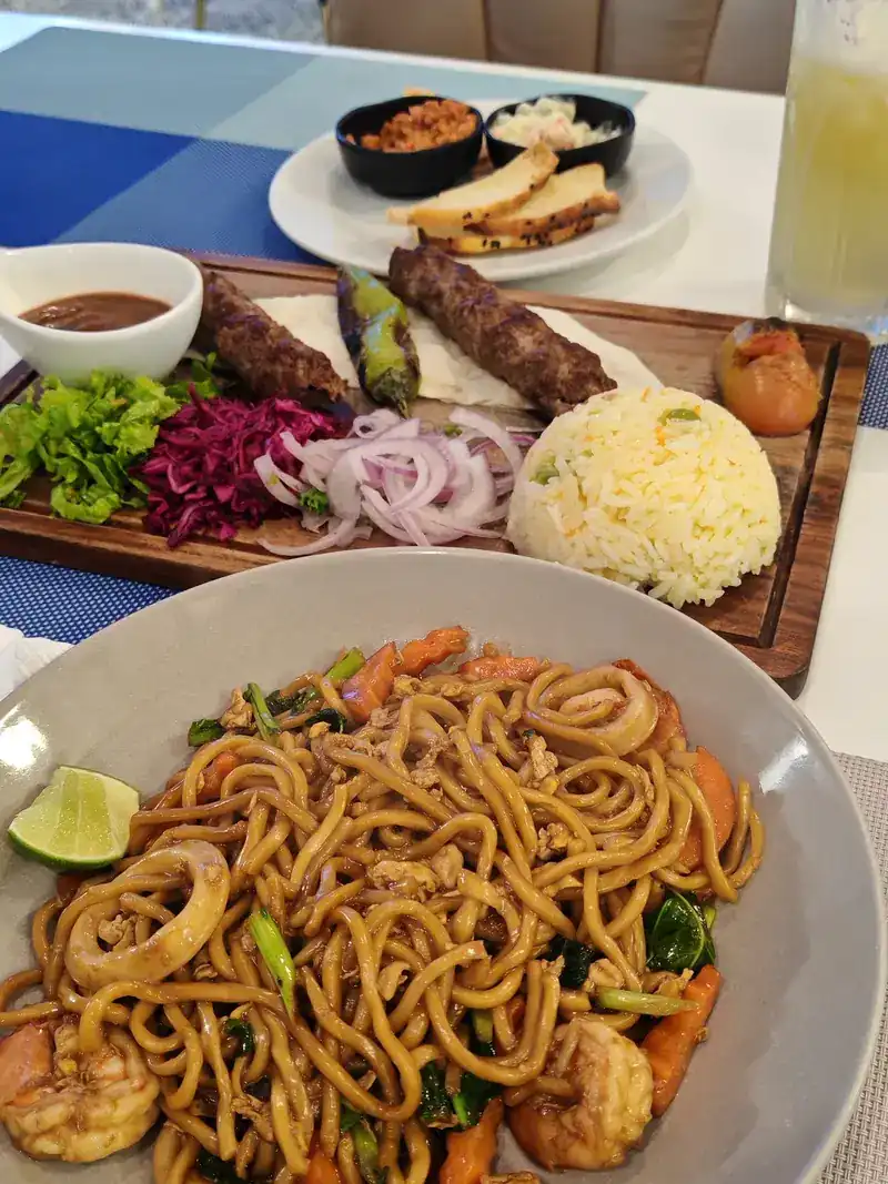 Adana kebab on a board and a bowl of stir-fried noodles with prawns and squid on the same table