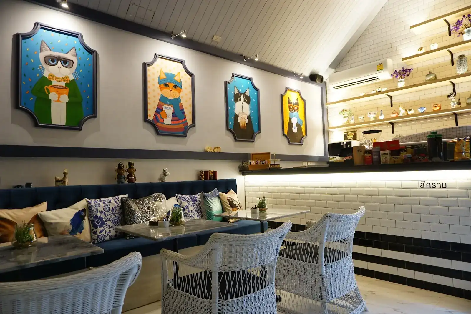 Inside Sea Calm Cafe: four painted cats in navy frames above a navy sofa with blue-and-white cushions, white wicker chairs and the white tiled counter