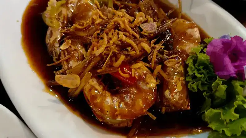 Prawns in a glossy dark sauce under a crisp fried topping, beside lettuce and a pink flower