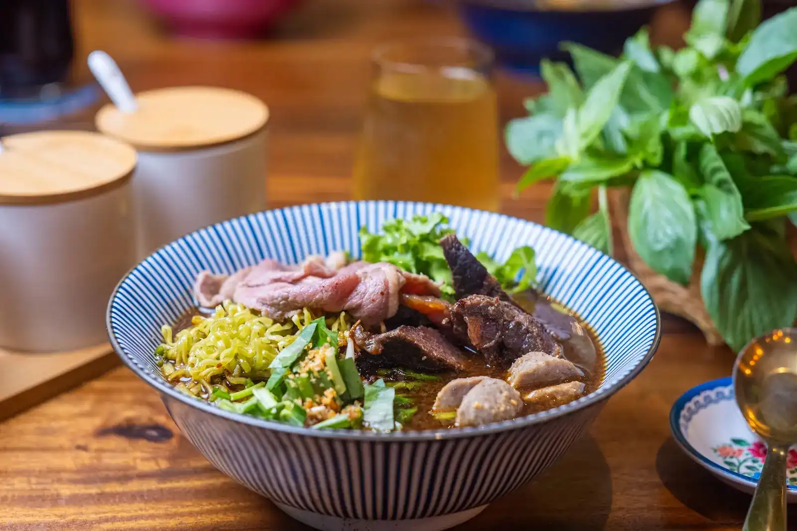 A bowl of beef noodles in a dark broth with sliced beef, braised beef and meatballs, in a blue-striped bowl beside a bunch of basil