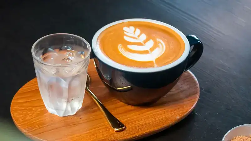 A latte with a leaf drawn in the foam, served on a wooden tray with a glass of water