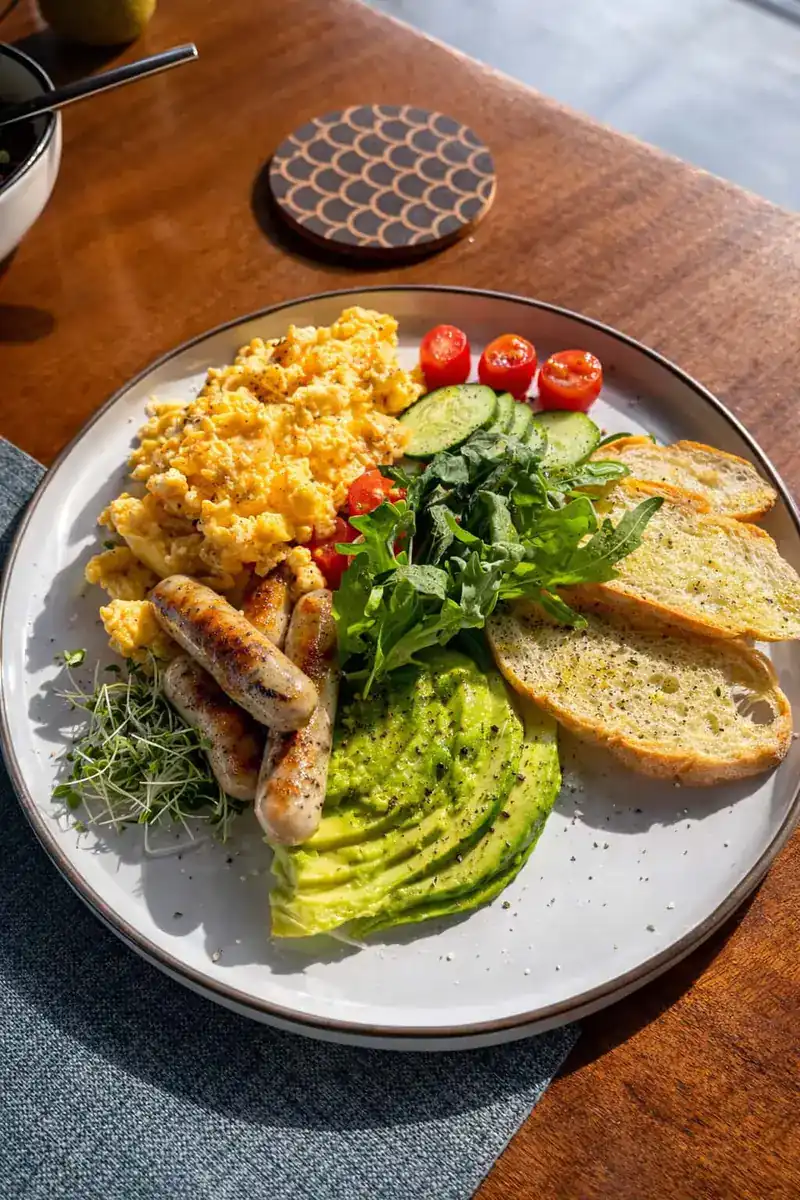 A plate of scrambled eggs, sausages, sliced avocado, salad and toast on a wooden table