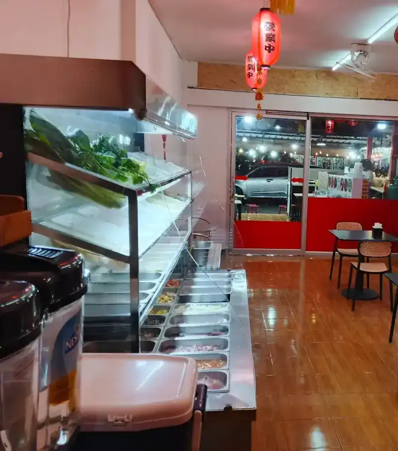 The open chiller inside the shop, greens on the top shelf and trays of ingredients below, under red lanterns