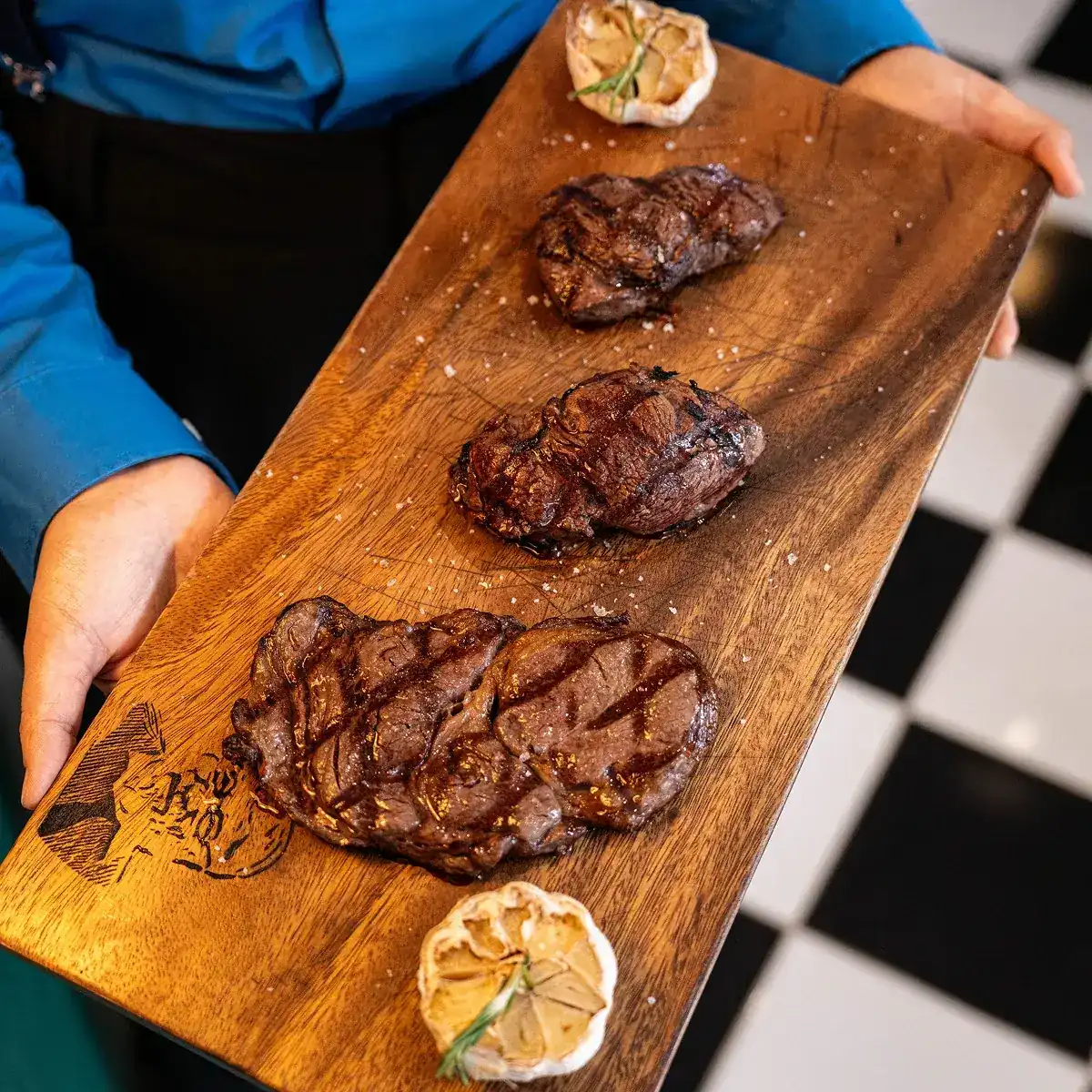 Three charcoal-grilled steaks with roasted garlic on a wooden board, carried to the table