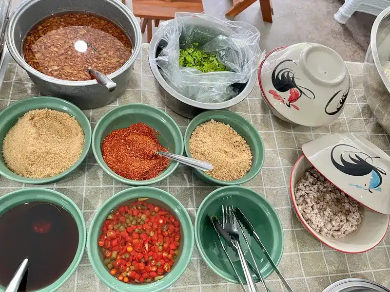 The condiment table: dried chilli, sugar, ground peanut and fresh chilli in green bowls, beside the rooster bowls