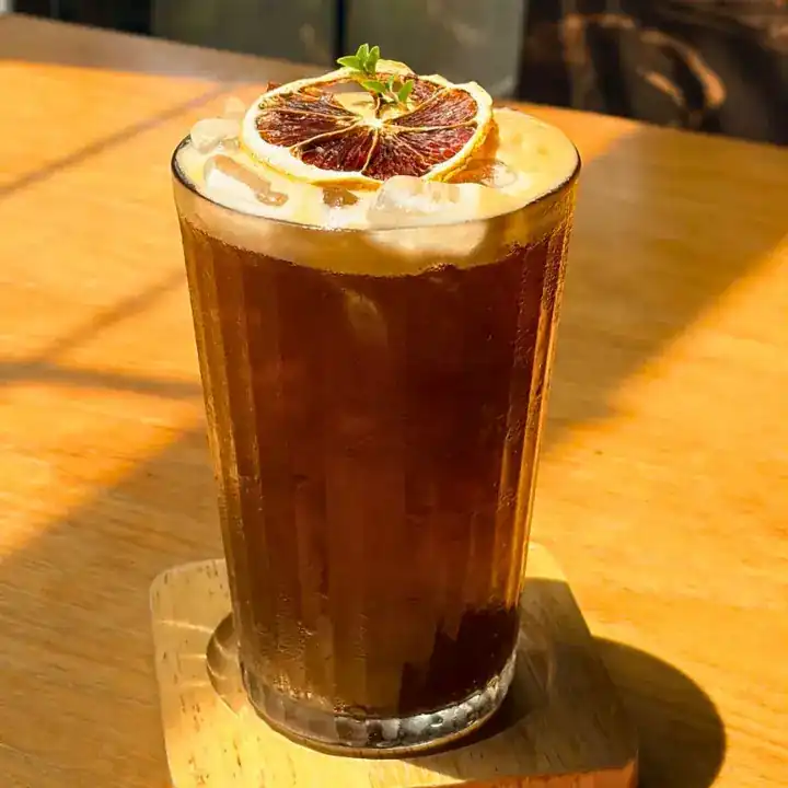 An iced drink with a creamy top and a dried orange slice, in the afternoon sun