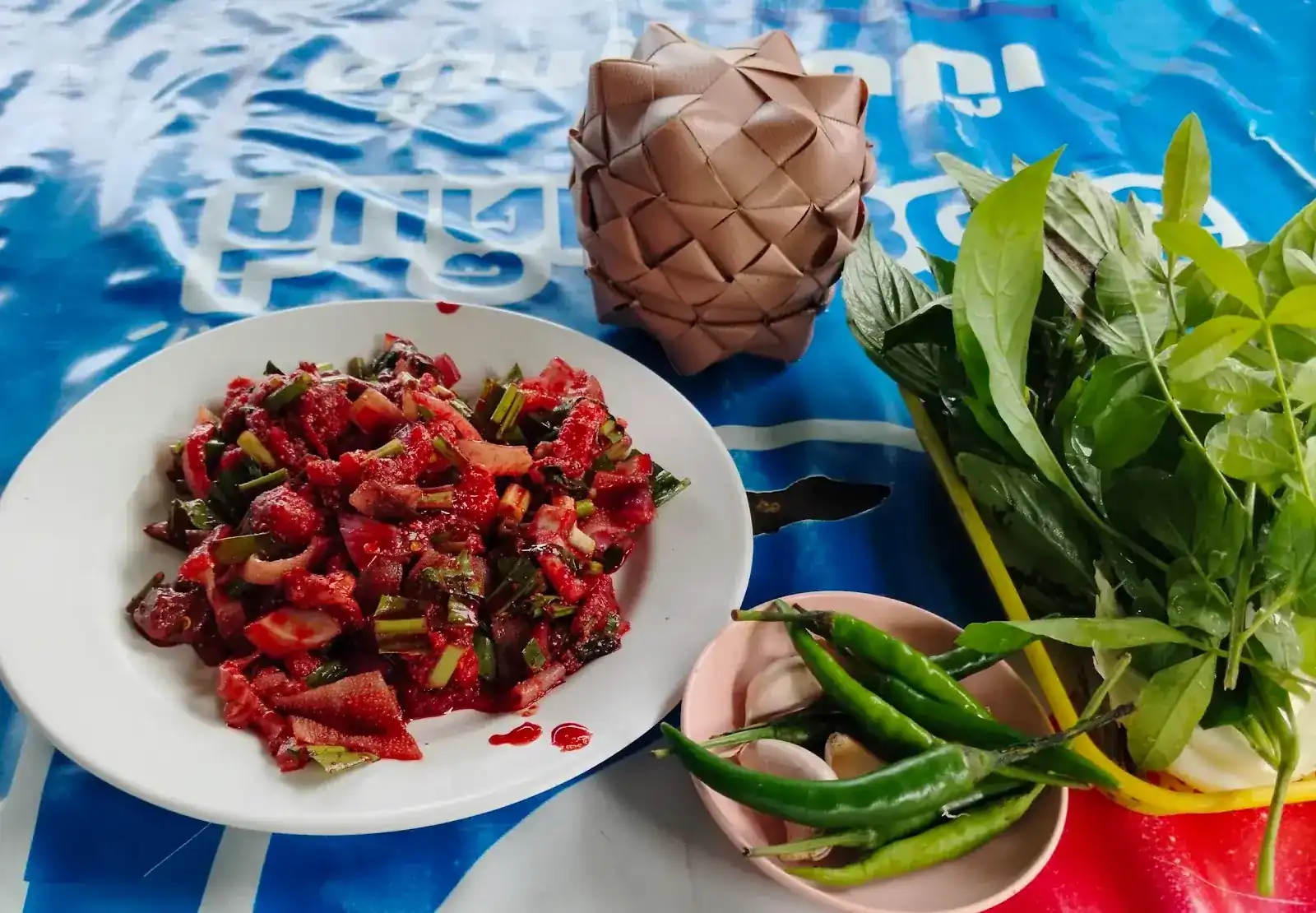 A red, chilli-dressed plate with spring onion, a woven leaf basket, fresh herbs, green chillies and garlic on the table
