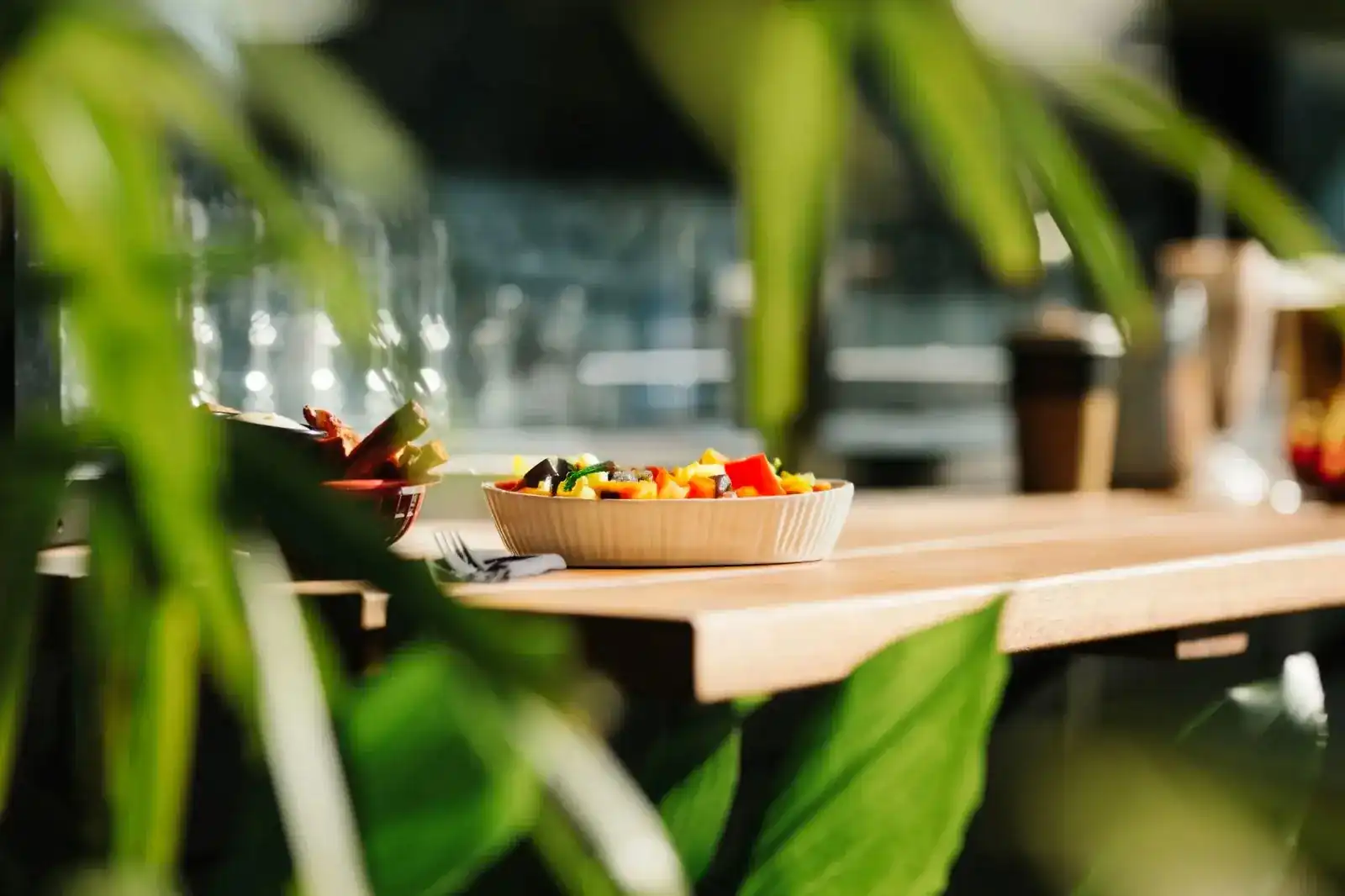 A bowl of fresh vegetables on the wooden counter, seen through green leaves