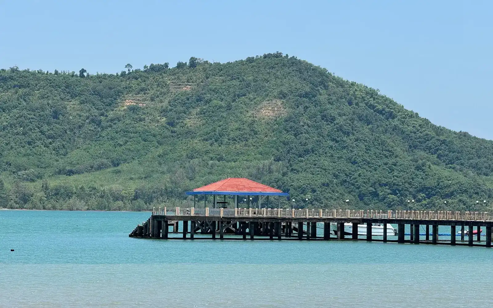 Turquoise water, a long pier with a red-roofed shelter and a green hill behind / น้ำทะเลสีเขียวคราม สะพานท่าเรือกับศาลาหลังคาแดง และเนินเขาเขียวด้านหลัง