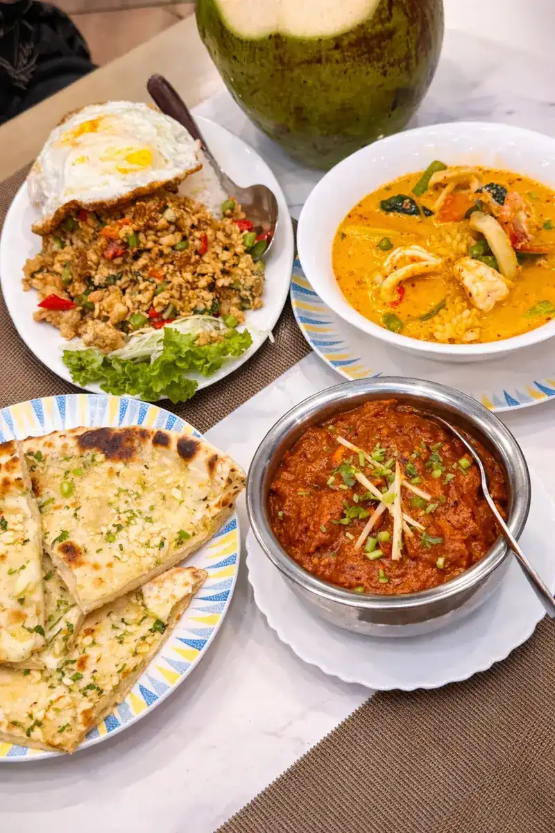 Thai and Indian dishes on one table: a stir-fry with a fried egg, a curry, naan and a fresh coconut