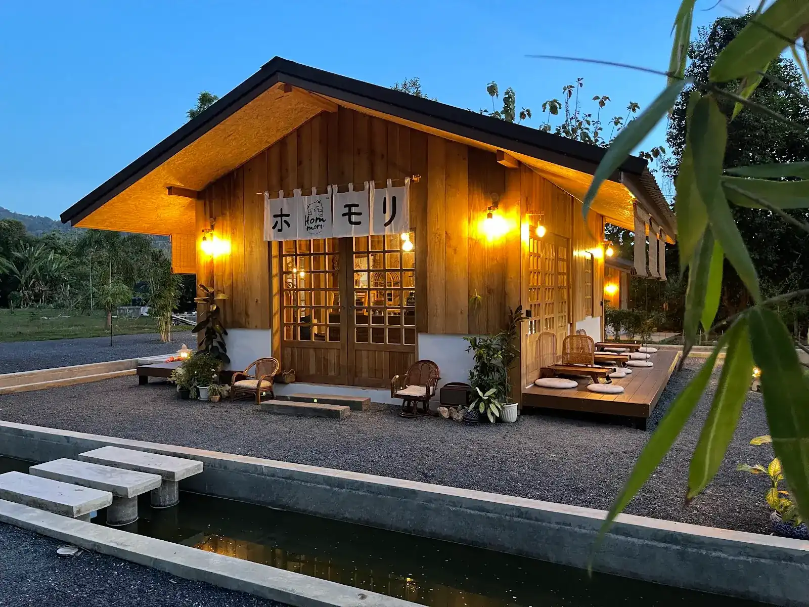Hommori's wooden house at dusk, lamps lit, with the ホモリ noren over the door and stepping stones across the water channel