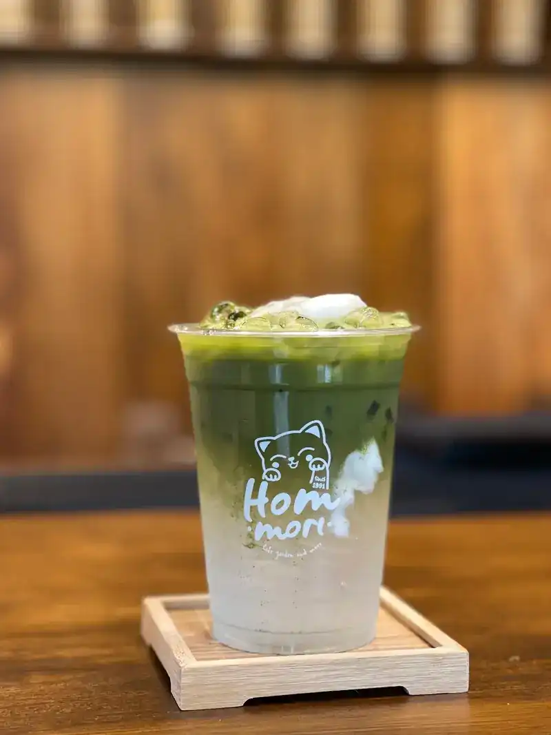 A layered iced matcha latte in a Hommori cup on a wooden coaster