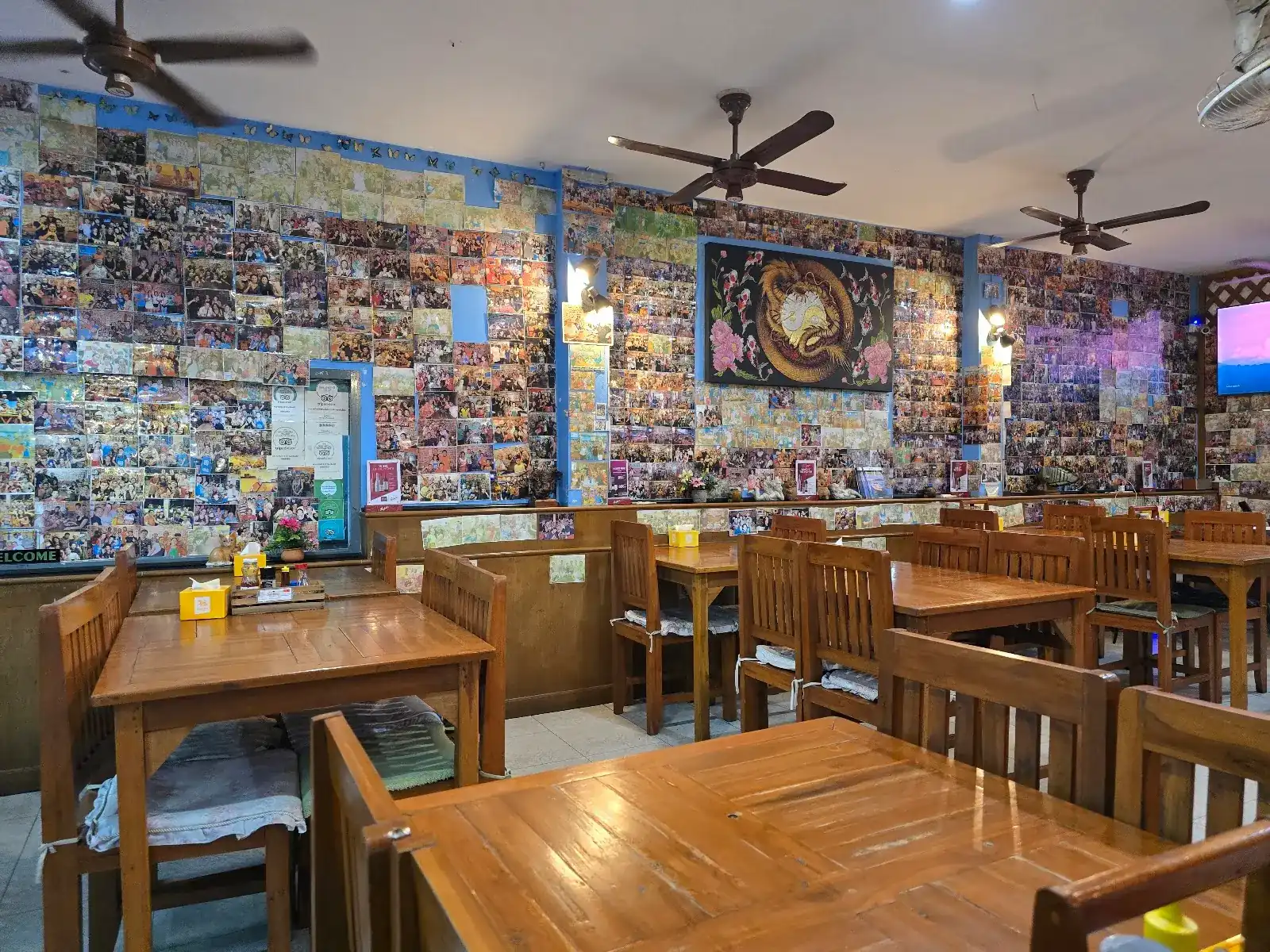 The dining room: wooden tables under ceiling fans, walls covered in photographs / ห้องอาหาร โต๊ะไม้ใต้พัดลมเพดาน ผนังติดรูปถ่ายเต็มห้อง