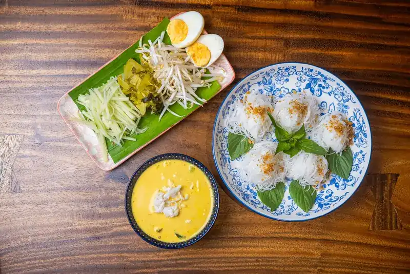 Khanom jeen seen from above: nests of fresh rice noodles with basil, a bowl of yellow curry, and boiled egg, bean sprouts and green papaya on a banana leaf
