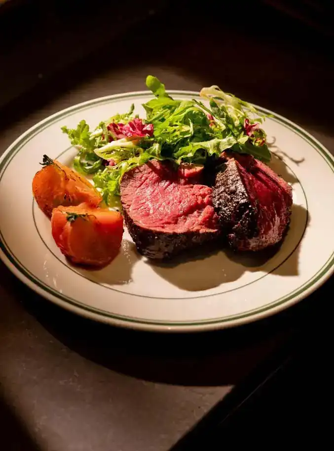 Tenderloin steak with leaves and roasted tomato on a green-rimmed plate, lit from the side