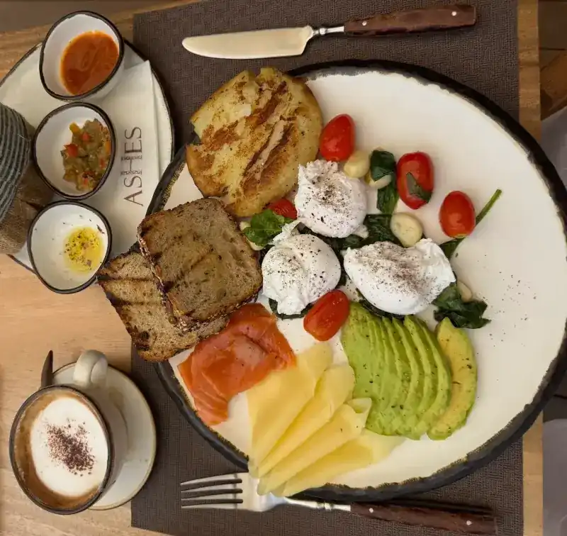 A breakfast plate with poached eggs, smoked salmon, avocado, cheese and toast, beside a cappuccino