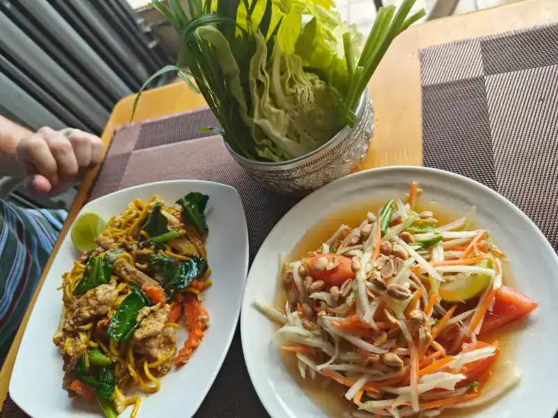 Stir-fried noodles with greens and a papaya salad topped with peanuts, beside a silver bowl of cabbage and fresh greens