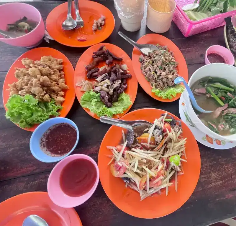 A table of Isan dishes on orange plates: papaya salad, larb, fried and grilled meat with lettuce, a soup with greens and bowls of dipping sauce