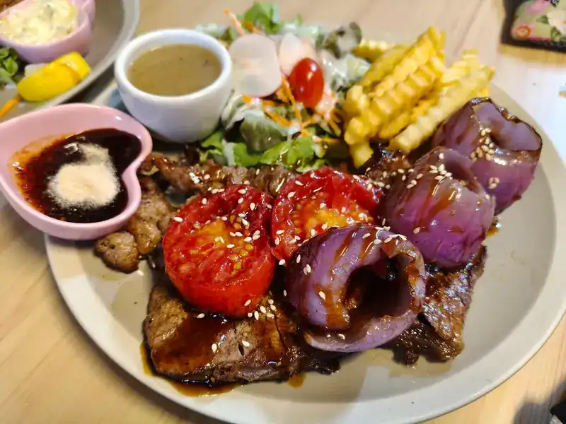 Steak glazed with sauce and sesame under grilled tomatoes and red onion, with salad, crinkle-cut fries and dipping sauce in a pink heart-shaped dish