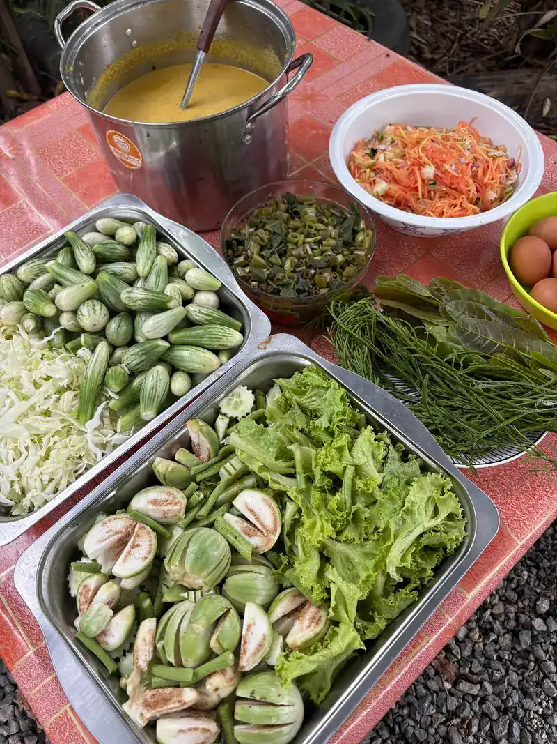 A pot of curry beside trays of fresh cucumbers, Thai eggplants, lettuce and herbs