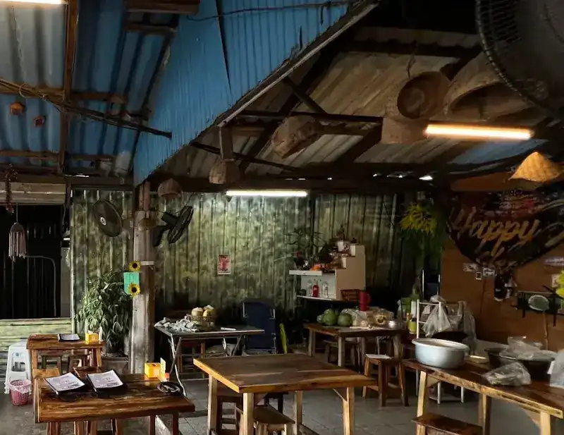 The open-sided shop under a tin roof, with wooden tables and stools / ร้านเปิดโล่งใต้หลังคาสังกะสี โต๊ะและเก้าอี้ไม้