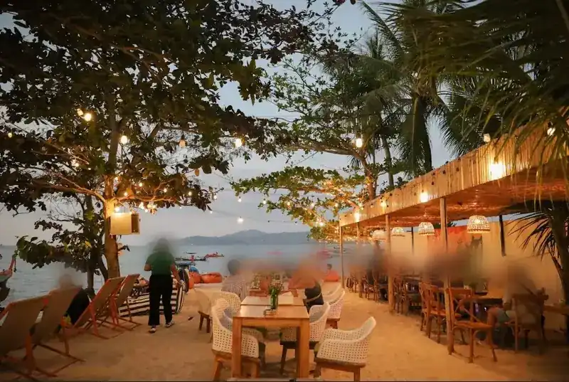 The beachfront terrace at dusk, string lights in the trees and tables on the sand