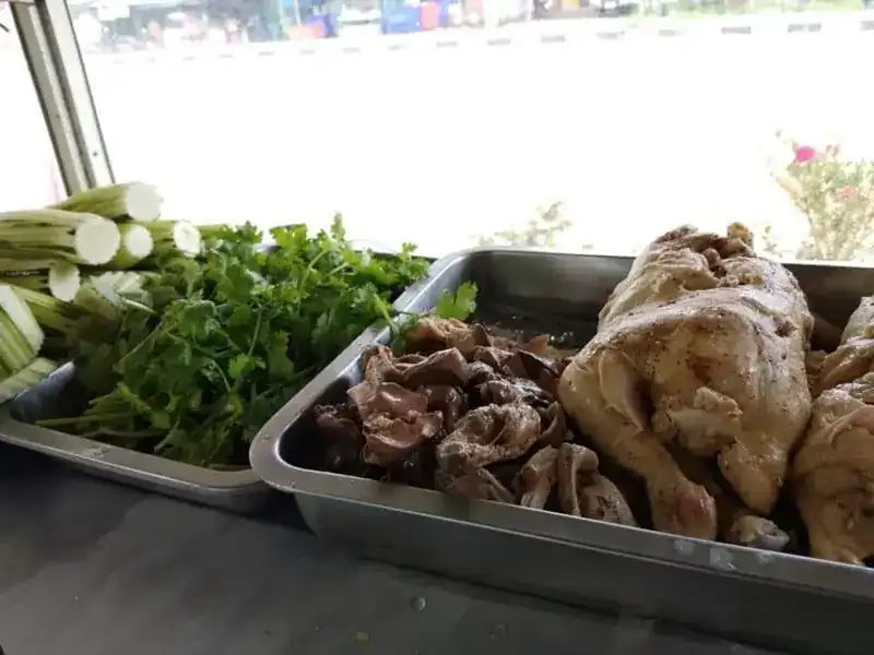 Steel trays of boiled chicken, chicken giblets, coriander and spring onion at the counter, with the road outside